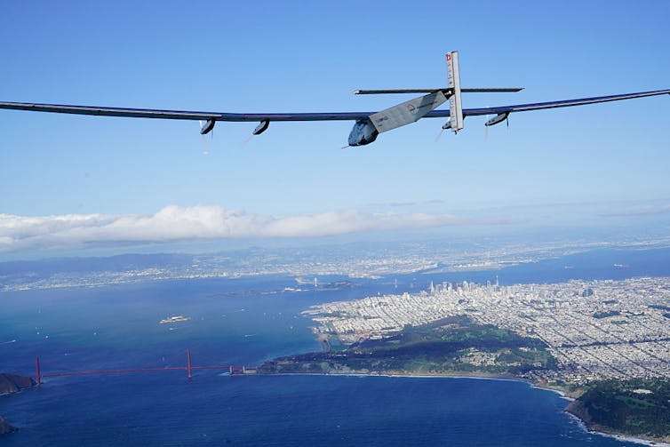 Futuristic looking plane with long wingspan flies over bay and city.