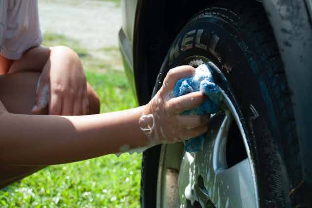 Hands of a person crouched down washing the wheel hub of a car, grass in the background.