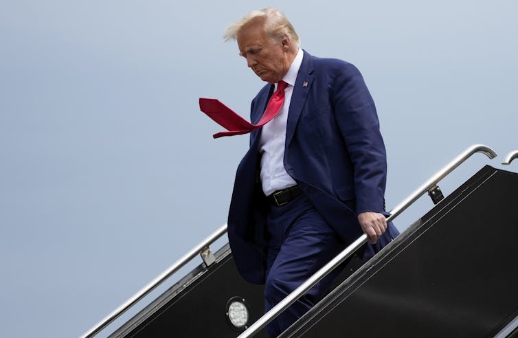 A blond man wearing a blue suit and a red tie steps out of an aircraft. His tie and hair blow in the wind.