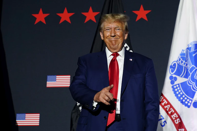 A tanned man with fluffy blond hair smiles while pointing at someone.