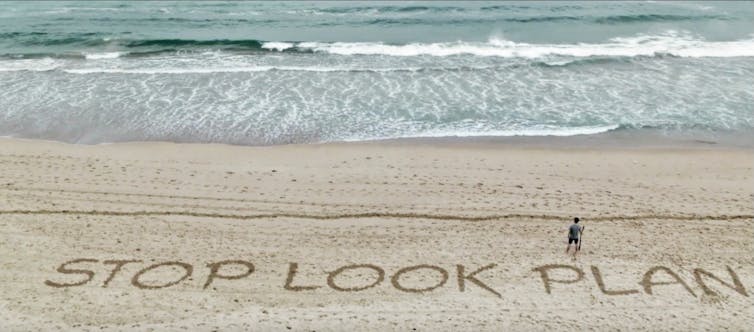 Person walking on a surf beach along a line with the words in the sand: Stop. Look. Plan.