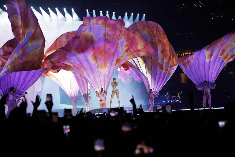 A singer on stage at a concert with a group of dancers with huge purple capes being blown upwards by wind machines.