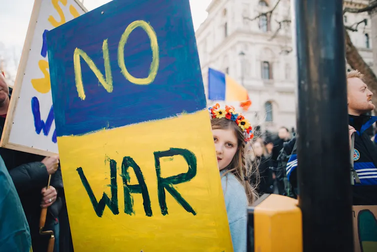 Child with flowers in hair holding sign with Ukrainian flag colours that says