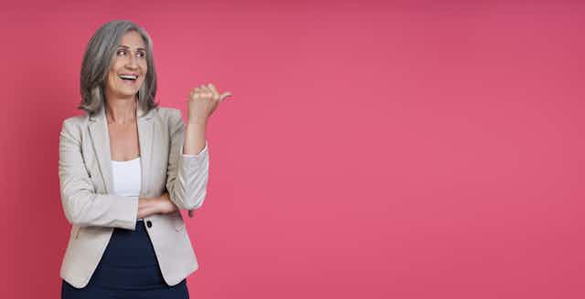 A grey-haired woman flicks her thumb up against a pink background