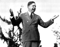 Un homme en costume-cravate fait un geste alors qu’il prononce un discours sur une photo en noir et blanc
