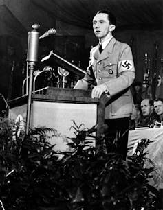 A black and white photo shows a thin dark-haired man in a Nazi uniform speaking into a microphone.