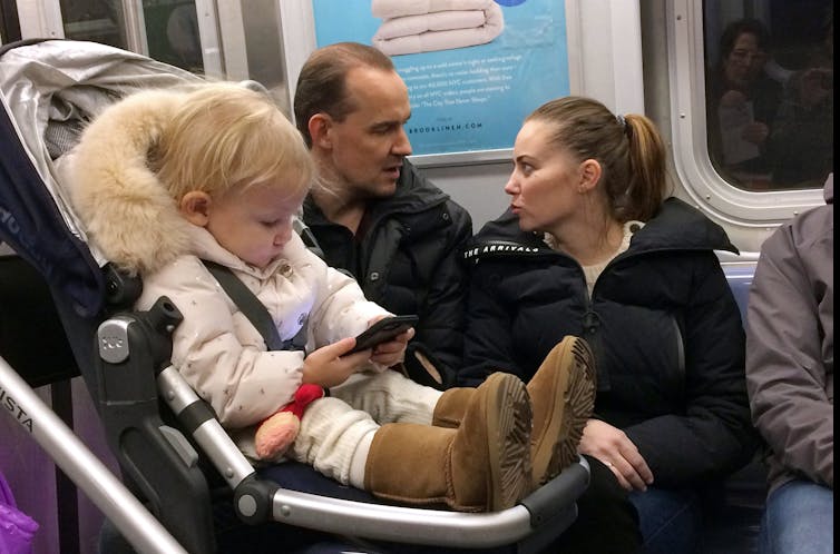A baby in a pram stares at a phone on a subway carriage.