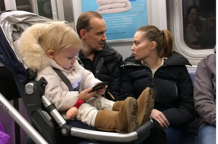 A baby in a pram stares at a phone on a subway carriage.