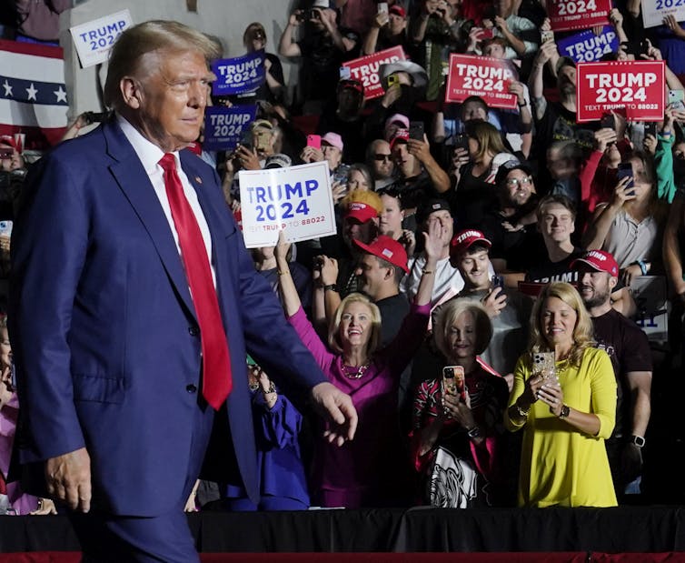 A blond man in a blue suit walks onto a stage as people smile and waves signs supporting him in the audience.