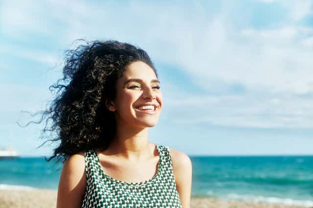 A smiling woman stands on a beach. She has dark curly hair.