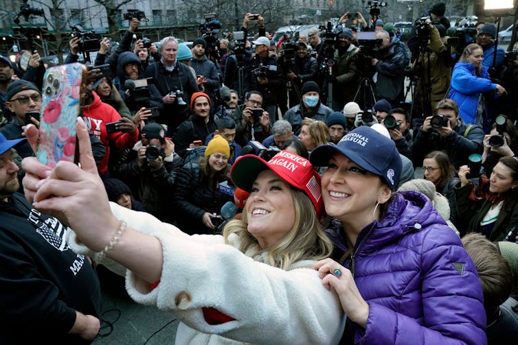 Two young white women wear Trump hats and take a selfie, in front of a crowd of people wearing winter clothing.