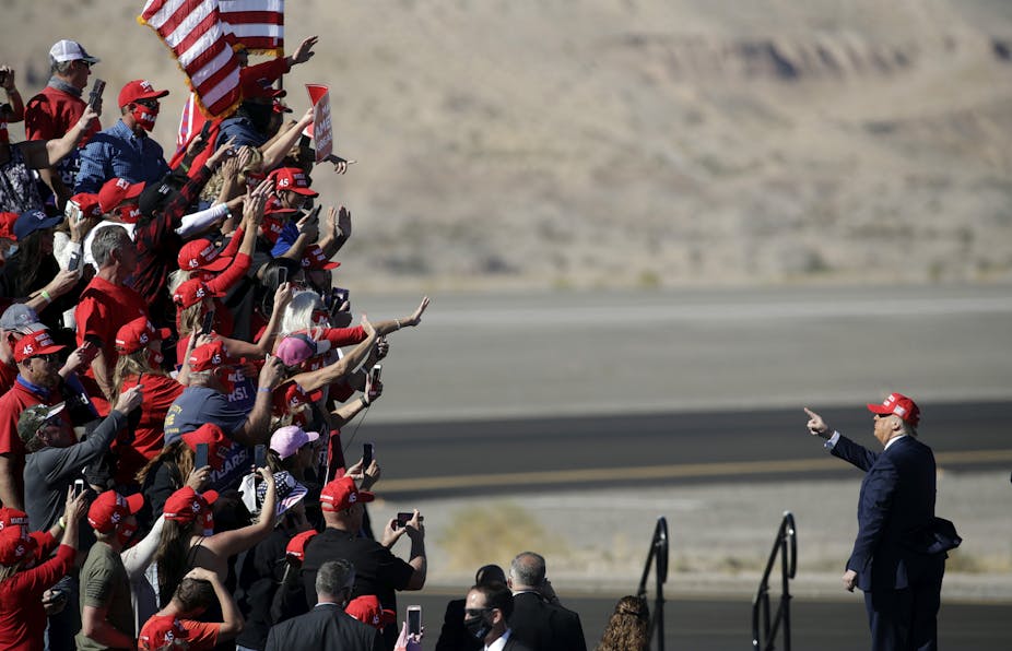 A crowd of people wear red and wave, facing Donald Trump in an arid looking outdoor space.