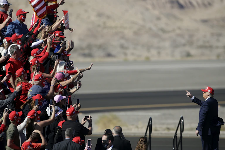 A crowd of people wear red and wave, facing Donald Trump in an arid looking outdoor space.