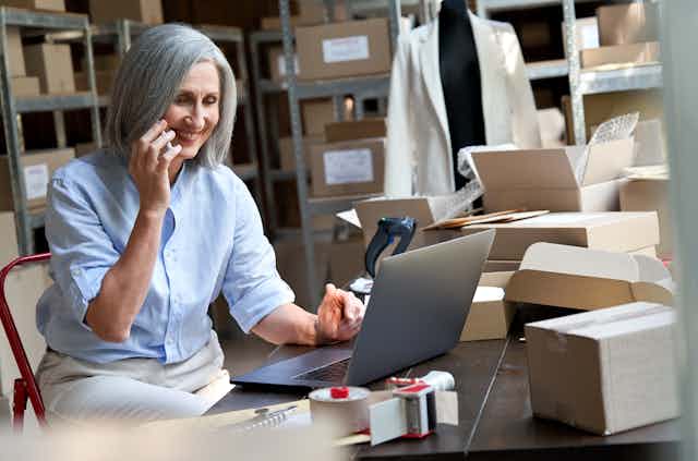 Woman with grey hair, smiling, on the phone, at a laptop, surrounded by boxes, shelves and papers, mannequin wearing jacket in the background.
