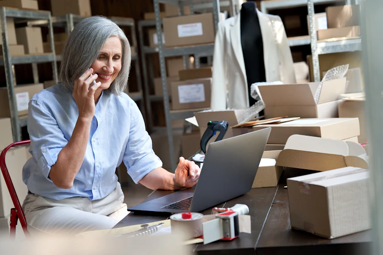 Woman with grey hair, smiling, on the phone, at a laptop, surrounded by boxes, shelves and papers, mannequin wearing jacket in the background.