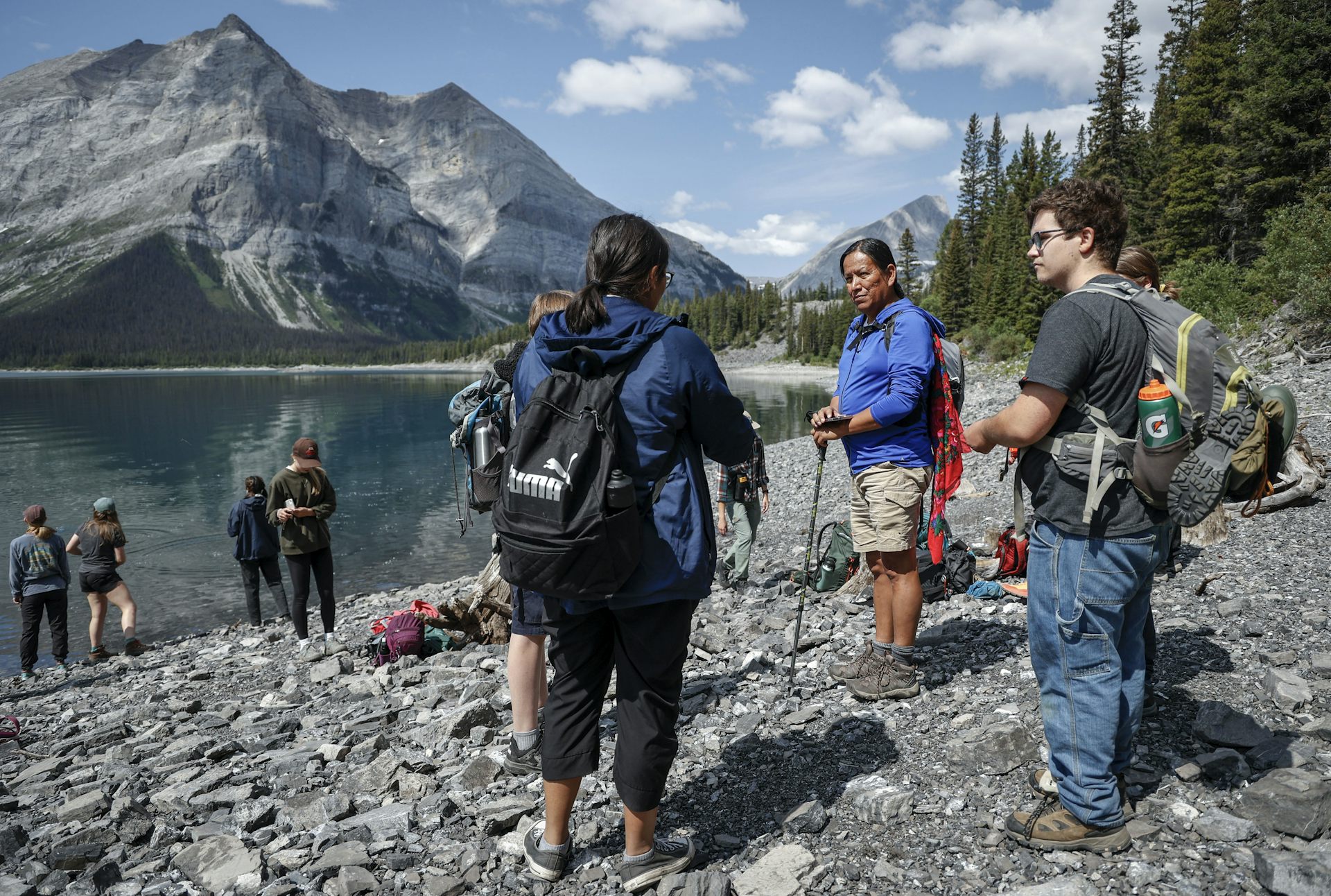 Indigenous Knowledge Keeper speaks with a group of people in front of a lake and mountains.