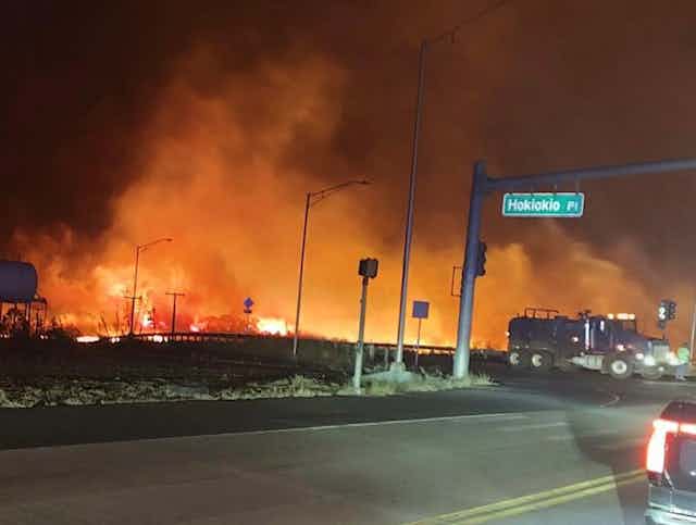 Flames line a road in the night as a water truck drives by.