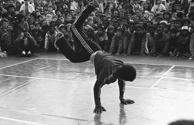 A black and white photo depicts a man breakdancing while a crowd look on.
