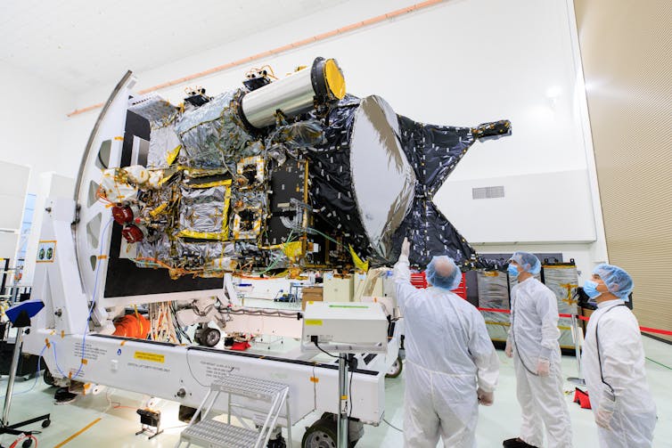 Technicians, inside a clean room and dressed in white garb, examine the Psyche spacecraft.