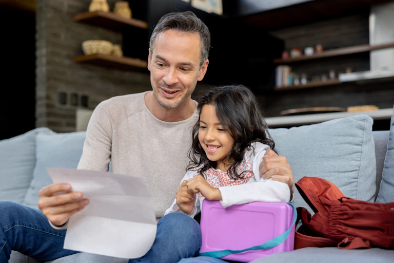 Father and young daughter sit on sofa looking at paper