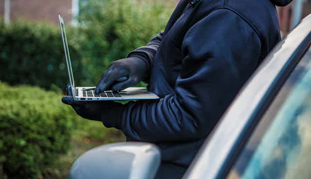 torso-only view of a man wearing gloves while using a laptop as he leans against a car