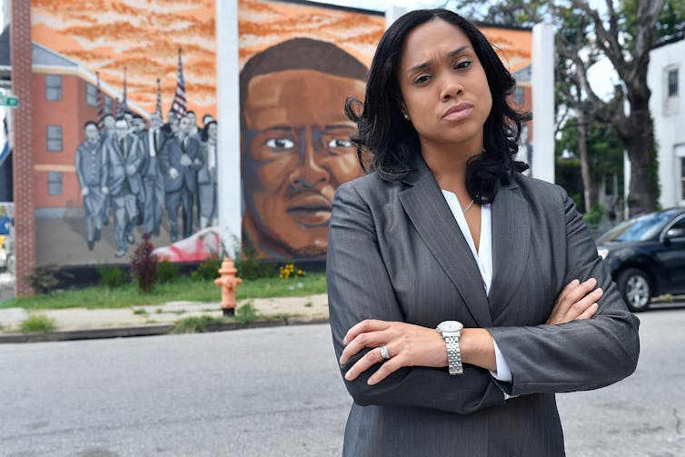 A Black woman dressed in a business suit stands in front of a mural with her arms crossed.