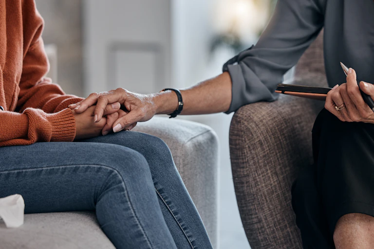 Two women whose faces are not visible hold hands as they sit in armchairs.
