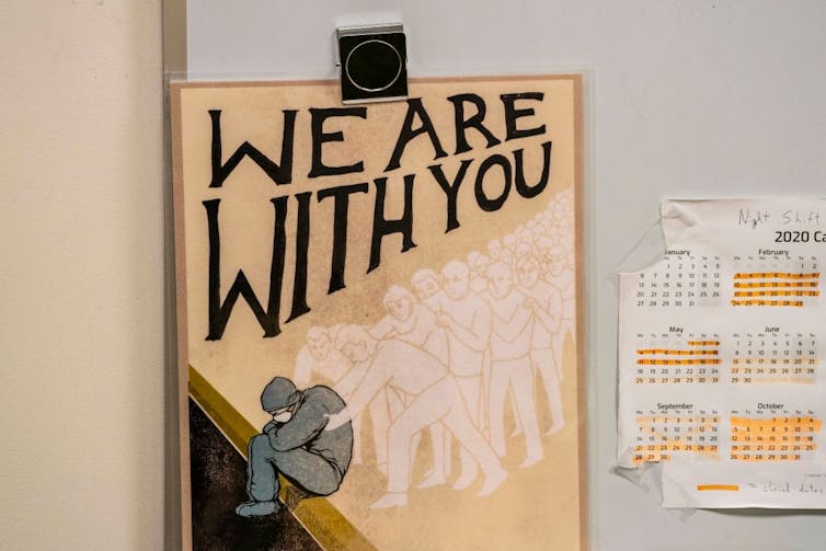 A poster that says 'We are with you,' with an illustration of someone sitting in scrubs as dozens of ghostly figures hold them.