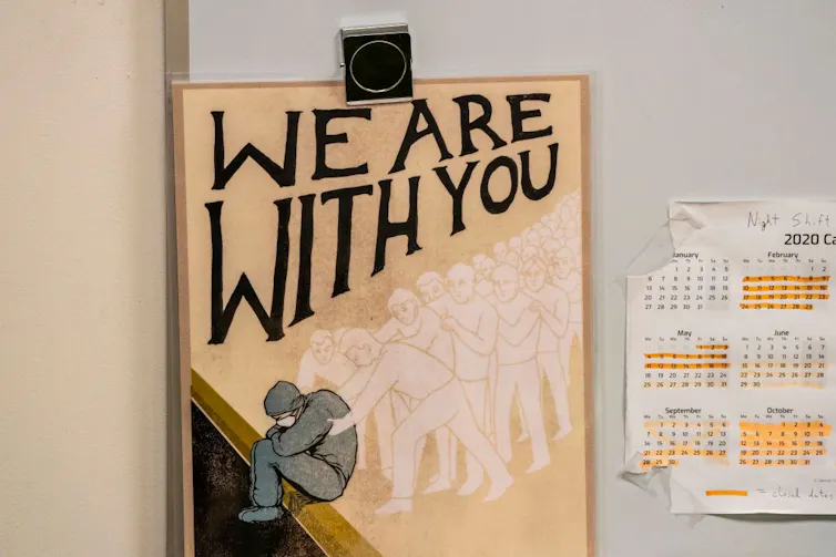 A poster that says 'We are with you,' with an illustration of someone sitting in scrubs as dozens of ghostly figures hold them.