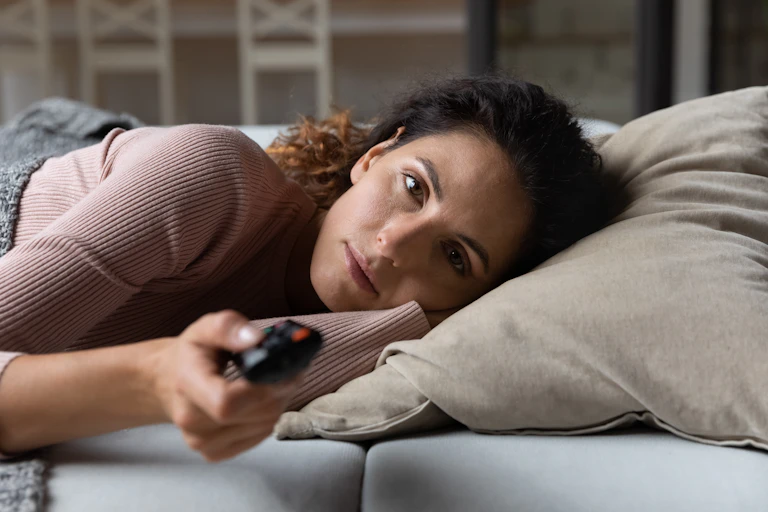A bored woman lays on her couch watching TV.