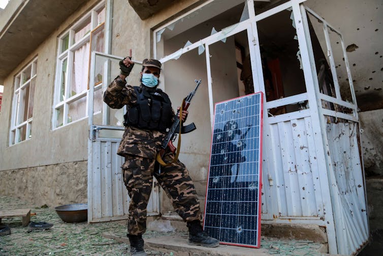 An armed Taliban fighter outside a house riddled with bullet holes and broken windows.