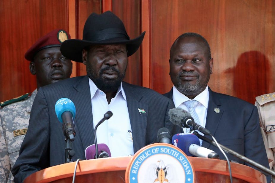 A man dressed in a shirt, blazer and a hat speaks at a podium written 'Republic of South Sudan' with microphones facing him. Another man in a suit stands behind him to his left, while a soldier in military fatigues stands at attention to his right