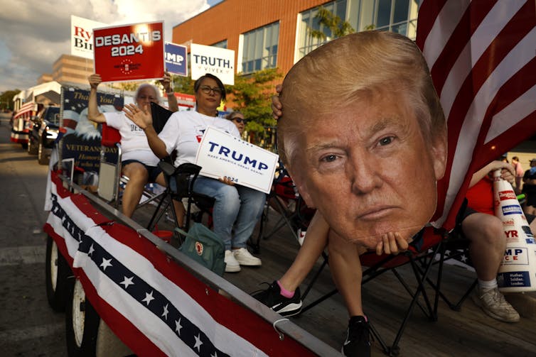 People wave Trump and DeSantis banners, along with a large picture of Trump's face, as they sit on top of a float with a red, white and blue star banner on the side of it.
