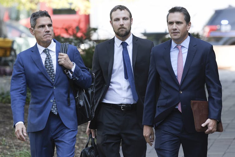 Three men in suits walk along a sidewalk.