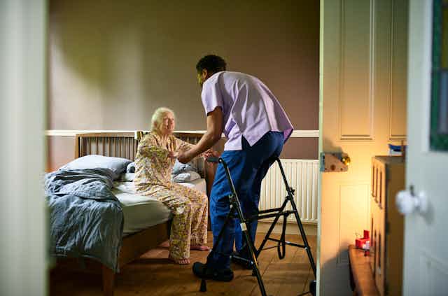 In a dimly-lit room, a male nurse helps an elderly woman sit up from her bed.