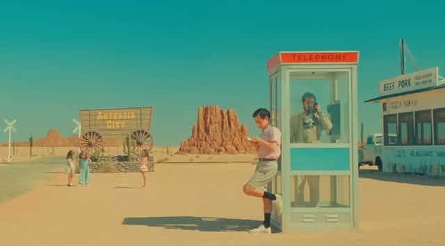 A scene from 'Asteroid City' with a man in a telephone booth and another leaning against it, in a stylized southwestern landscape