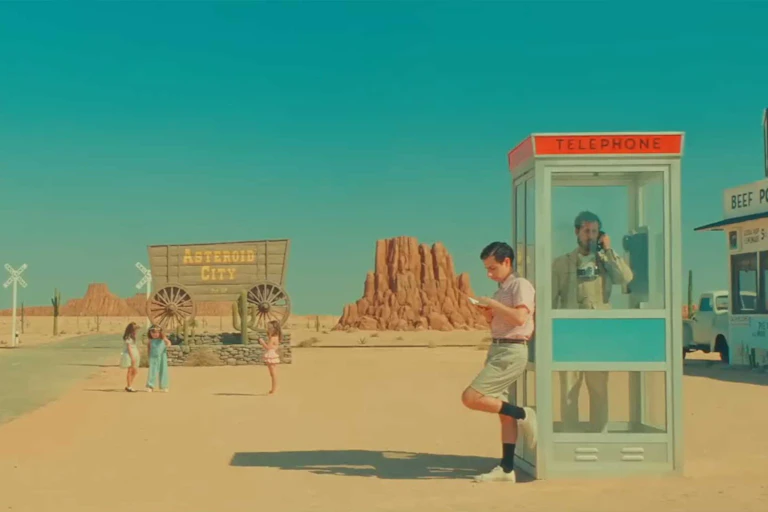 A scene from 'Asteroid City' with a man in a telephone booth and another leaning against it, in a stylized southwestern landscape