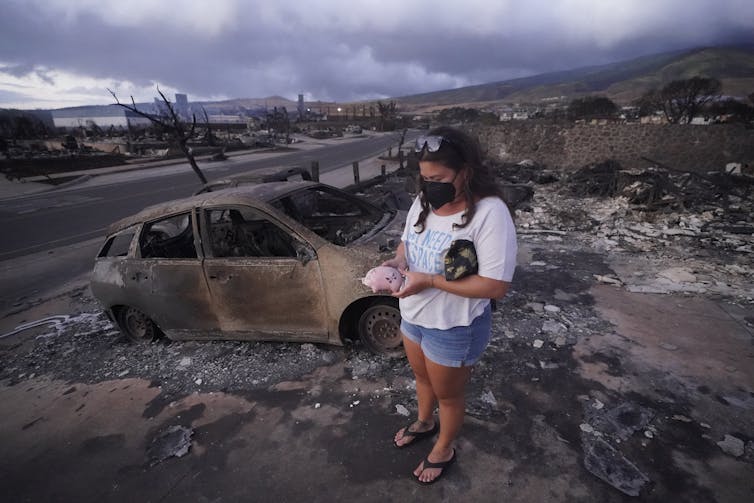 A woman in flipflops, shorts and a T-shirt holds a small pink pig-shaped piggy bank, surrounded by a burned car and charred landscape. The ocean is in the background.