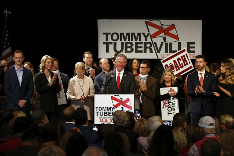 A man in a dark suit, white shirt and red tie, talks on a stage surrounded by a crowd.