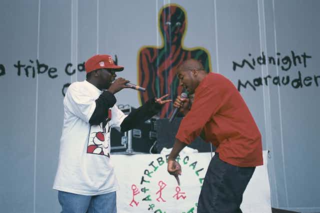 Two men in long-sleeve t-shirts sing into microphones in front of a wall with graffiti.