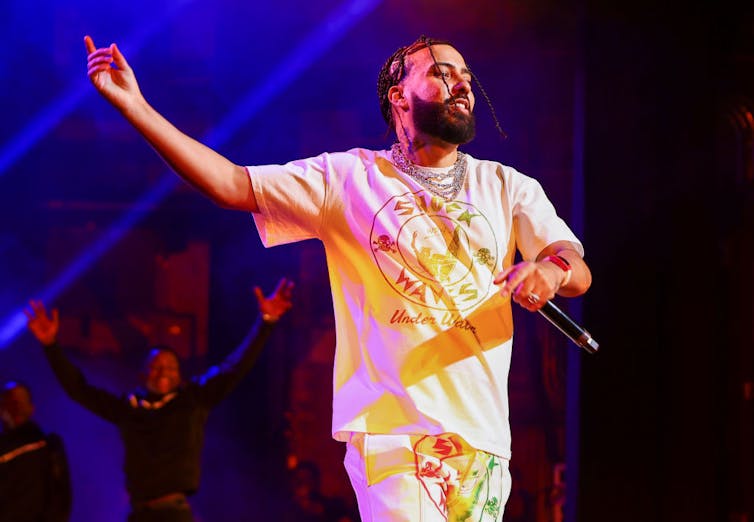 A man with braided hair in a white t-shirt with neon patterns on it gestures and holds a microphone on stage.