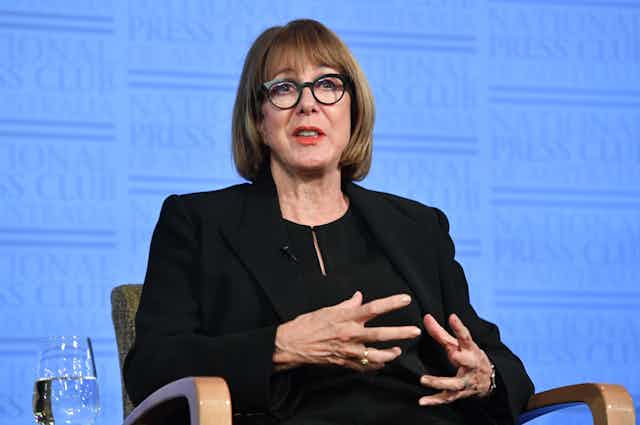 woman wearing glasses is sitting and speaking to an audience
