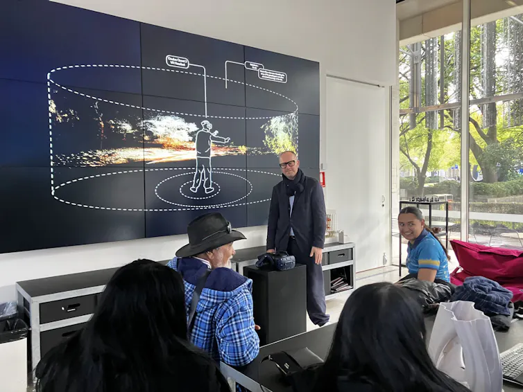 Three people are in front of a large screen which shows an image of a person wearing a VR headset while walking through a created landscape.