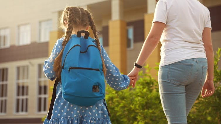A mother leads her child by the hand toward a building. The child is wearing a backpack.