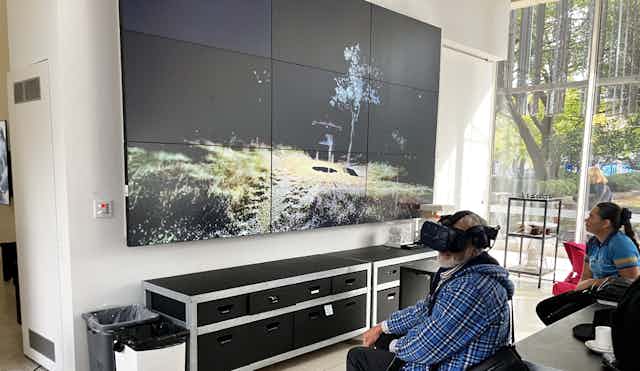 Two people sit in front of a large screen. One of the people wears a VR headset. The screen shows an area outside.