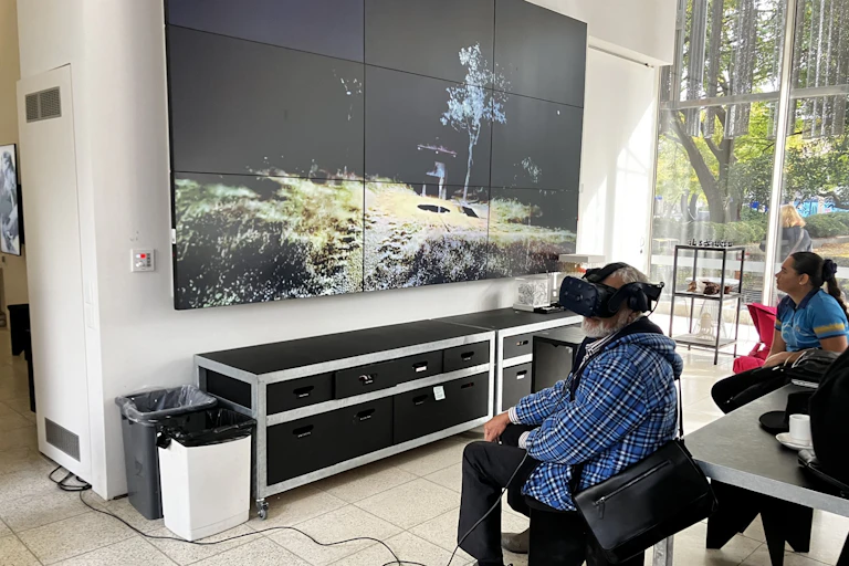 Two people sit in front of a large screen. One of the people wears a VR headset. The screen shows an area outside.
