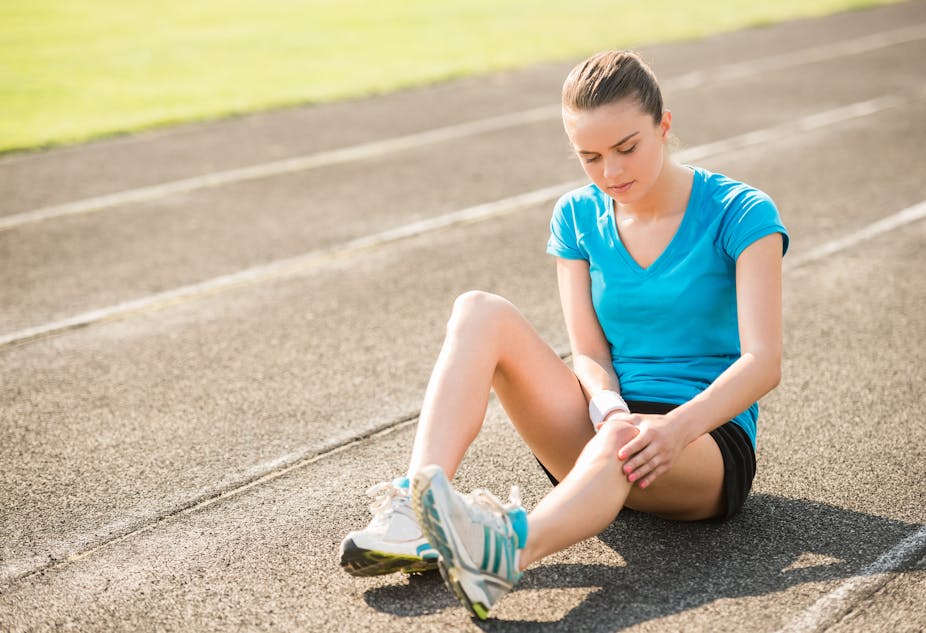 A young women sits on an outdoor running track, holding her knee in pain.