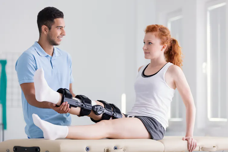 A young woman wearing a knee brace for her ACL performs a leg lift with the help of her male physiotherapist.