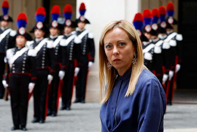 Giorgia Meloni looking to camera while standing in front of Italian troops standing to attention.