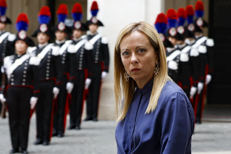 Giorgia Meloni looking to camera while standing in front of Italian troops standing to attention.